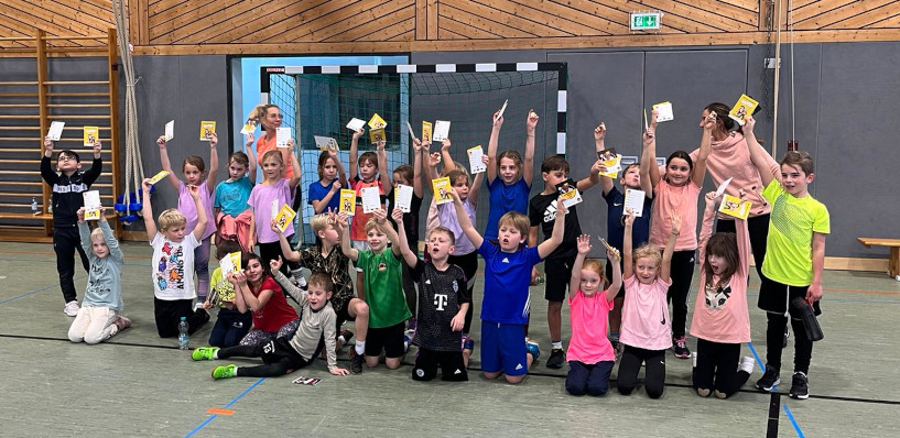 Alle teilnehmenden Zweitklässer stehen oder knien vor einem Handballtor zusammen um fotografiert zu werden. Einige Helfer haben sich links und rechts dahinter gestellt. Alle strecken die Arme hoch und halten das offizielle Handball-Spielabzeichen des DHB – den Hanniball-Pass in der Hand.