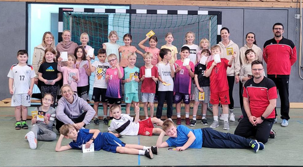 In der Nendinger Sporthalle haben sich vor einem Handballtor alle Kinder die beim HVW-Grundschulaktionstag dabei waren aufgestellt für ein gemeinsames Foto. In der letzten Reihe stehen die Kinder auf einer Bank. In der Reihe davor zeigen einige Kinder stehend voller Stolz das offizielle Handball-Spielabzeichen des DHB – den Hanniball-Pass. In der vordersten Reihe liegen einige Kinder seitlich vor den anderen. Die erwachsenen Helfer und Organisatoren stehen jeweils links und rechts neben den Kindern.