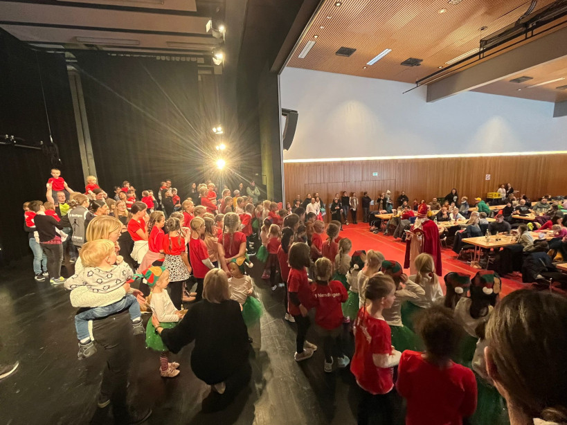 In der Mehrzweckhalle der Donau-Hallen steht vor der Bühne der Nikolaus. Auf der Bühne stehen ganz viele Kinder und hören dem Nikolaus gespannt zu. Im Saal sieht man an Tischreihen die Eltern und Angehörigen.