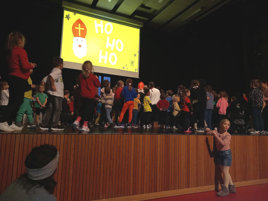 In der Nendinger Donau-Hallen in der Mehrzweckhalle auf der Bühne stehen viele Kinder und hören dem Nikolaus der in der Mitte steht aufmerksam zu. Im Hintergrund ist die Leinwand ausgefahren und darauf sieht man einen Nikolaus und den Text: Ho,Ho,Ho