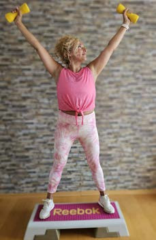 Die Kursleiterin steht für ein Foto in einer Wohnung vor einer Wand auf einem Stepbrett mit kleinen gelben Hanteln jeweils in den Händen und streckt die Arme nach oben.