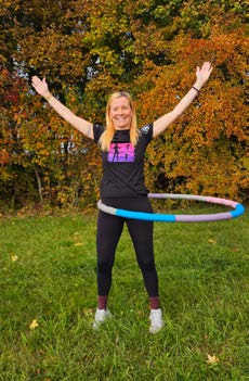 In einem Garten auf dem Rasen steht Doreen mit ihrem Hula Hoop-Reifen um der Taillie. Ihre Arme streckt sie dabei seitlich noch oben.