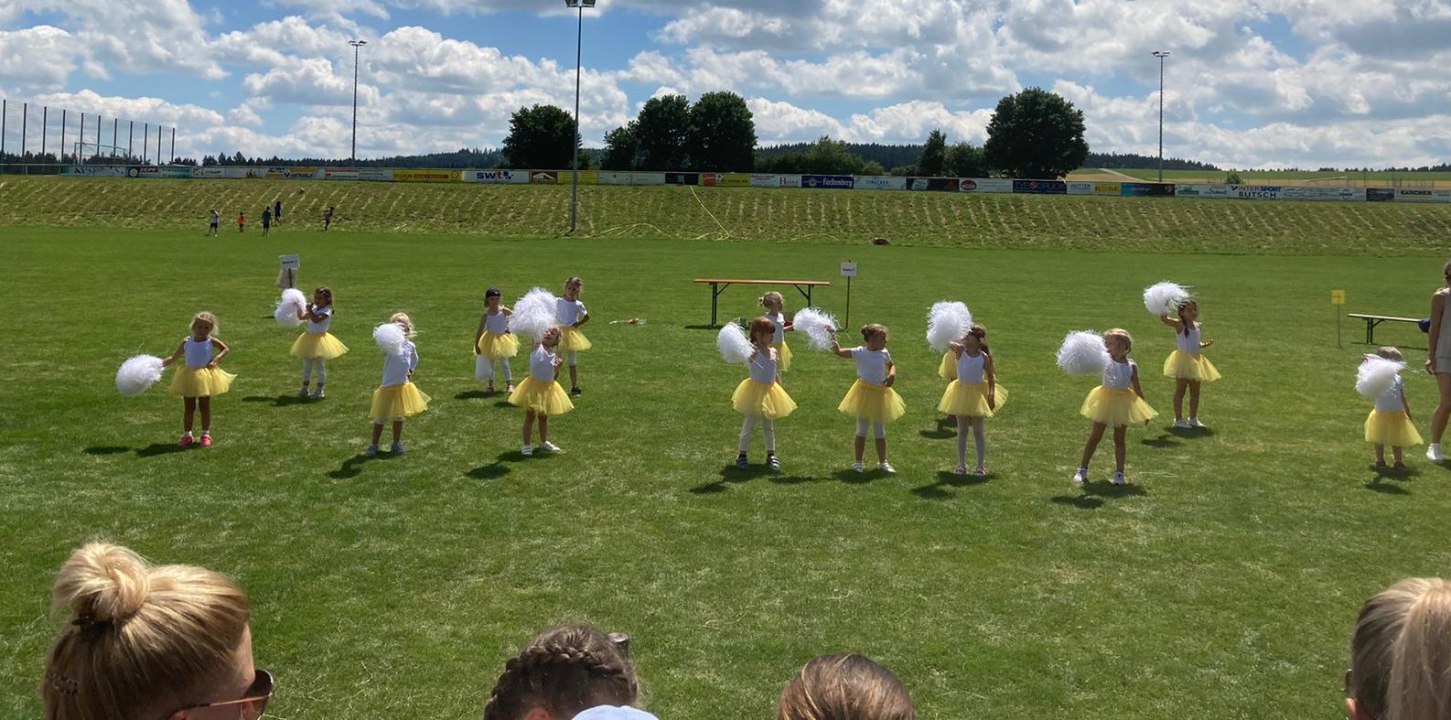Auf dem Fußballspielfeld auf dem Häldele stehen viele Kinder der Gruppe Tanzmäuse mit ihren tollem Kostümen und den weißen Pom-Poms auf ihren Positionen und tanzen nach lauter Musik. Im Vordergrund stehen Erwachsene und schauen sich den niedlichen Tanz genau an.