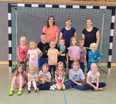 Für ein Gruppenfoto haben sich 14 Kinder in einer Sporthalle zusammengestellt vor einem Handballtor. Die drei Trainerinnen stehen dahinter und lächeln mit in die Kamera.